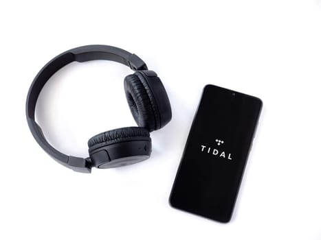 Lod, Israel - July 16,2023: Smartphone with TIDAL Music app launch screen and wireless headphones on white background. Top view flat lay with copy space.