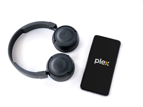 Lod, Israel - July 16,2023: Smartphone with Plex app launch screen and wireless headphones on white background. Top view flat lay with copy space.