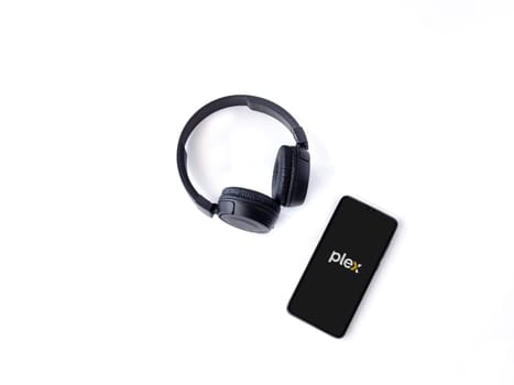Lod, Israel - July 16,2023: Smartphone with Plex app launch screen and wireless headphones on white background. Top view flat lay with copy space.