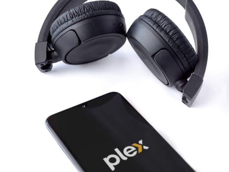 Lod, Israel - July 16,2023: Smartphone with Plex app launch screen and wireless headphones on white background. Top view flat lay with copy space.
