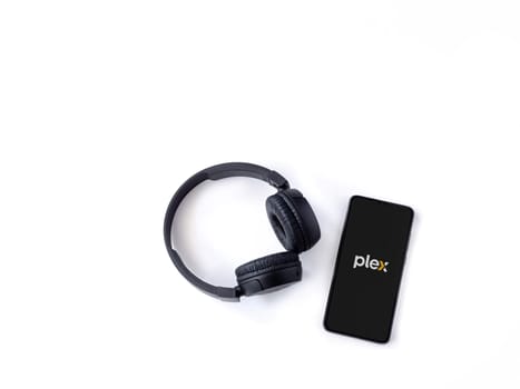 Lod, Israel - July 16,2023: Smartphone with Plex app launch screen and wireless headphones on white background. Top view flat lay with copy space.