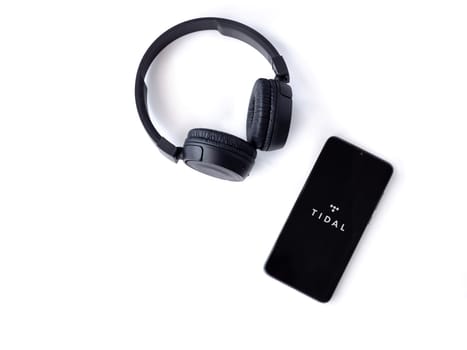 Lod, Israel - July 16,2023: Smartphone with TIDAL Music app launch screen and wireless headphones on white background. Top view flat lay with copy space.