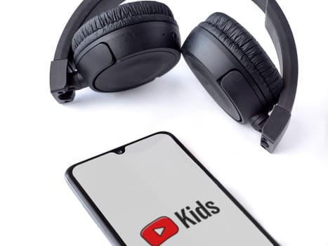Lod, Israel - July 16,2023: Smartphone with YouTube Kids app launch screen and wireless headphones on white background. Top view flat lay with copy space.