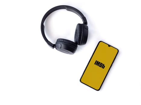 Lod, Israel - July 16,2023: Smartphone with IDMb app launch screen and wireless headphones on white background. Top view flat lay with copy space.