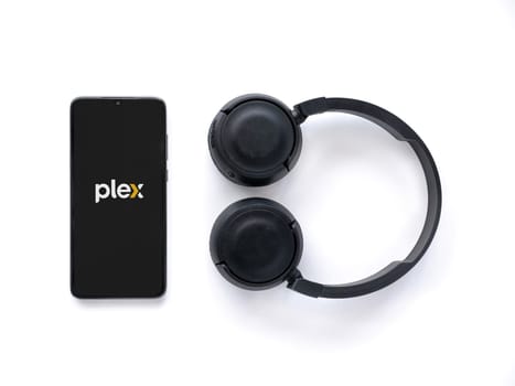 Lod, Israel - July 16,2023: Smartphone with Plex app launch screen and wireless headphones on white background. Top view flat lay with copy space.