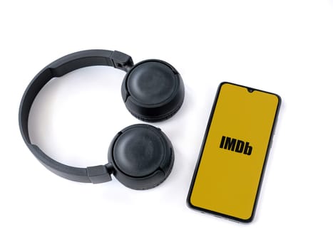 Lod, Israel - July 16,2023: Smartphone with IDMb app launch screen and wireless headphones on white background. Top view flat lay with copy space.