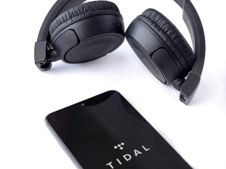 Lod, Israel - July 16,2023: Smartphone with TIDAL Music app launch screen and wireless headphones on white background. Top view flat lay with copy space.