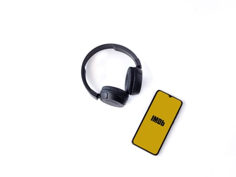 Lod, Israel - July 16,2023: Smartphone with IDMb app launch screen and wireless headphones on white background. Top view flat lay with copy space.