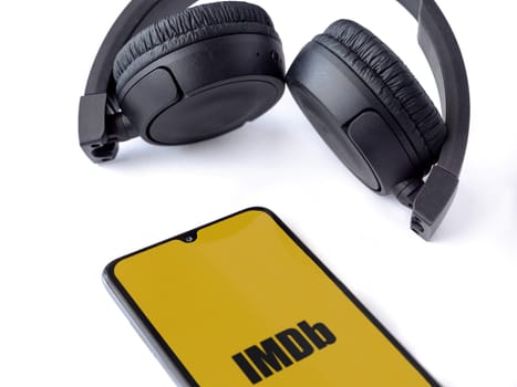 Lod, Israel - July 16,2023: Smartphone with IDMb app launch screen and wireless headphones on white background. Top view flat lay with copy space.