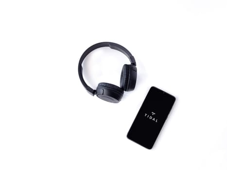 Lod, Israel - July 16,2023: Smartphone with TIDAL Music app launch screen and wireless headphones on white background. Top view flat lay with copy space.