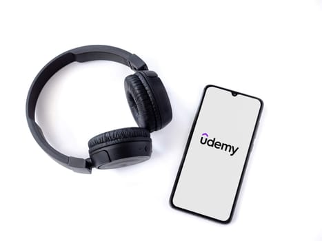 Lod, Israel - July 16,2023: Smartphone with Udemy app launch screen and wireless headphones on white background. Top view flat lay with copy space.
