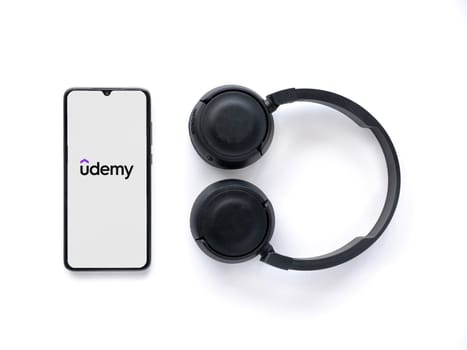 Lod, Israel - July 16,2023: Smartphone with Udemy app launch screen and wireless headphones on white background. Top view flat lay with copy space.