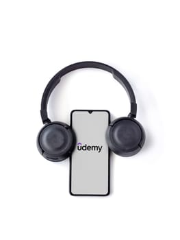 Lod, Israel - July 16,2023: Smartphone with Udemy app launch screen and wireless headphones on white background. Top view flat lay with copy space.