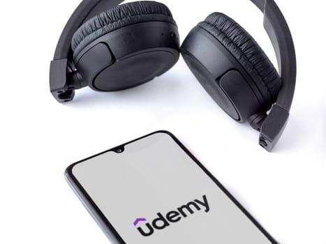 Lod, Israel - July 16,2023: Smartphone with Udemy app launch screen and wireless headphones on white background. Top view flat lay with copy space.