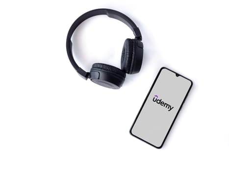 Lod, Israel - July 16,2023: Smartphone with Udemy app launch screen and wireless headphones on white background. Top view flat lay with copy space.