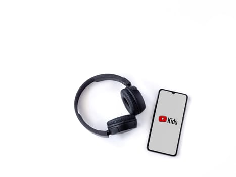 Lod, Israel - July 16,2023: Smartphone with YouTube Kids app launch screen and wireless headphones on white background. Top view flat lay with copy space.