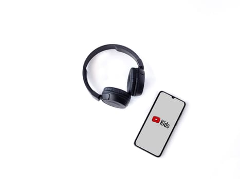 Lod, Israel - July 16,2023: Smartphone with YouTube Kids app launch screen and wireless headphones on white background. Top view flat lay with copy space.