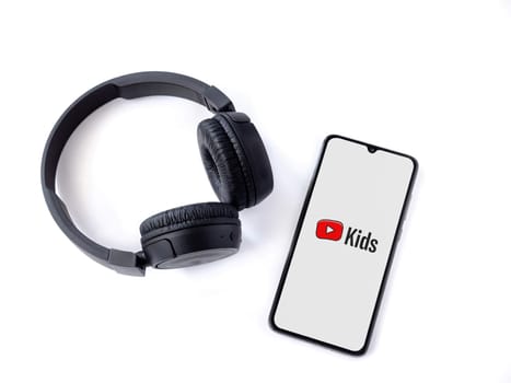 Lod, Israel - July 16,2023: Smartphone with YouTube Kids app launch screen and wireless headphones on white background. Top view flat lay with copy space.