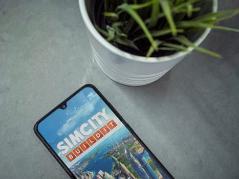 Lod, Israel - July 16,2023: Modern workspace with smartphone with SimCity BuildIt app launch screen on marble background. Close up top view flat lay.