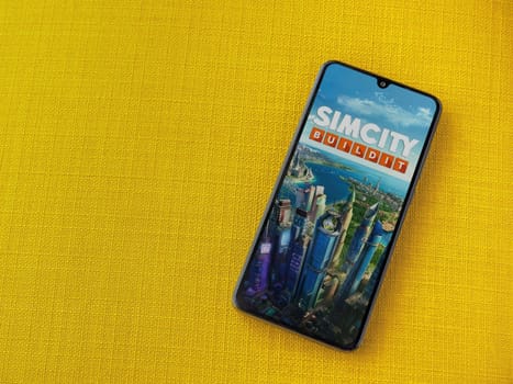 Lod, Israel - July 16,2023: SimCity BuildIt app launch screen on smartphone on yellow fabric background. Top view flat lay with copy space.