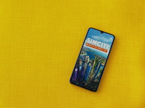 Lod, Israel - July 16,2023: SimCity BuildIt app launch screen on smartphone on yellow fabric background. Top view flat lay with copy space.