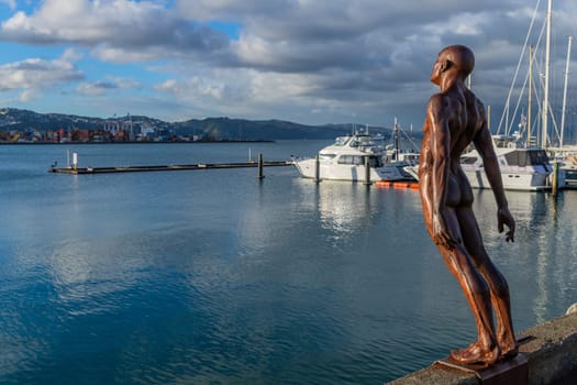 Wellington, New Zealand - May 13 2023 Statue of naked man leaning into wind on city waterfront with commercial skyline behind installed in 2007 by sculpture Max Patte.