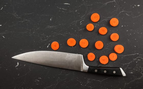 Top down view, chefs knife and small circles of sliced carrot on black marble like working board.