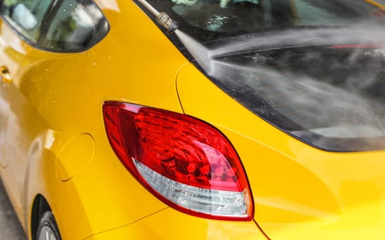 Rear window and red back light of yellow  car washed in self serve carwash, jet water spraying on glass.