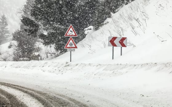 Sharp curve on road, with road signs (careful skid, snow) during heavy snowstorm blizzard in winter. Dangerous driving conditions.