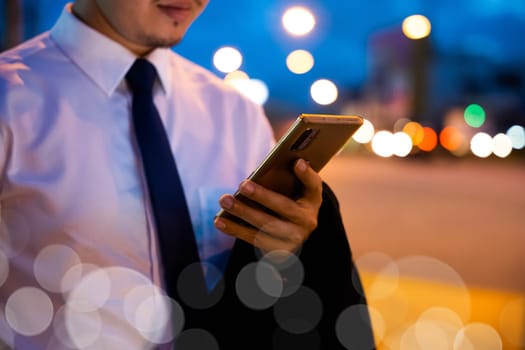 Portrait Asian businessman typing an sms message via smartphone after work near office at night city street, bokeh lights, young man walking and chatting with friends at social networks outdoor