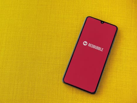 Lod, Israel - July 16,2023: Redbubble app launch screen on smartphone on yellow fabric background. Top view flat lay with copy space.