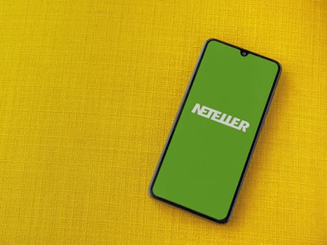 Lod, Israel - July 16,2023: NETELLER app launch screen on smartphone on yellow fabric background. Top view flat lay with copy space.