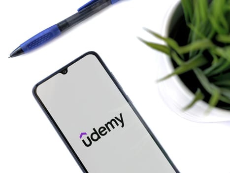 Lod, Israel - July 16,2023: Modern workspace with smartphone with Udemy app launch screen on white background. Close up top view flat lay.