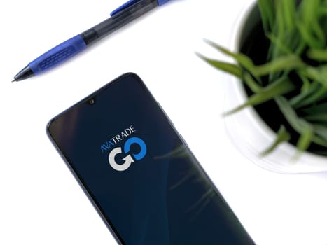 Lod, Israel - July 16,2023: Modern workspace with smartphone with Avatrade app launch screen on white background. Close up top view flat lay.