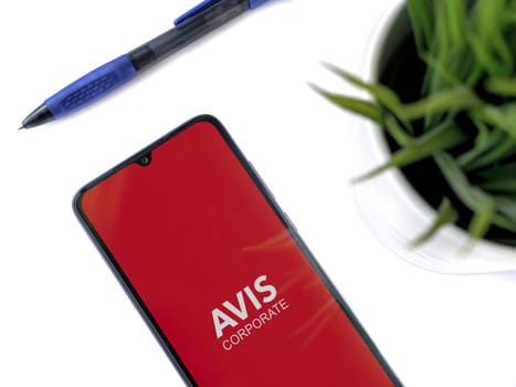 Lod, Israel - July 16,2023: Modern workspace with smartphone with Avis Corporate app launch screen on white background. Close up top view flat lay.