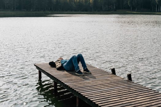 A man enjoys a sunny autumn day, resting lying on a wooden boat dock on a lake outside the city.
