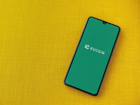 Lod, Israel - July 16,2023: KuCoin app launch screen on smartphone on yellow fabric background. Top view flat lay with copy space.