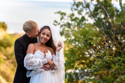 Newlyweds hug against the backdrop of a beautiful landscape, the guy kisses the girl who is looking into the frame