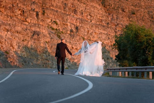 Happy newlyweds walk along a mountain asphalt road from the camera