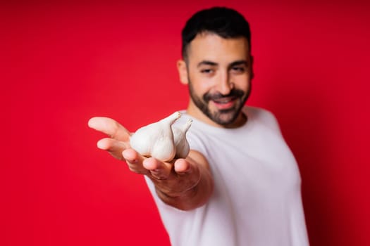 Young bearded man in a white shirt is holding garlics with smile, isolated over red background