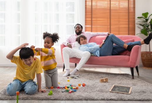 Curly little kindergarten girl teasing cute boy while playing wooden blocks together on floor in living room. Couple parent resting on couch while father scolding daugther and mother watching children.