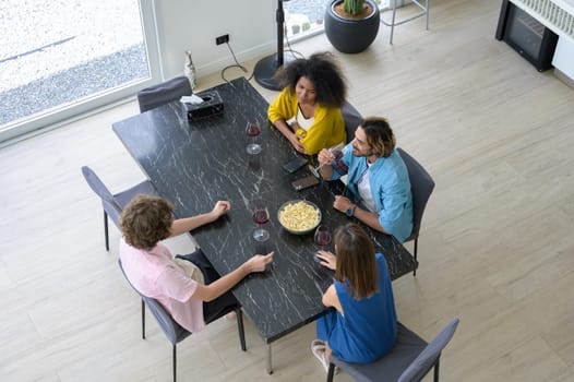 Multiracial group of happy young people sitting around table, drinking red wine with popcorn and having conversation together at home.