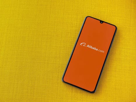 Lod, Israel - July 16,2023: Alibaba.com app launch screen on smartphone on yellow fabric background. Top view flat lay with copy space.