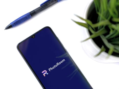 Lod, Israel - July 16,2023: Modern workspace with smartphone with PhotoRoom Studio Photo Editor app launch screen on white background. Close up top view flat lay.