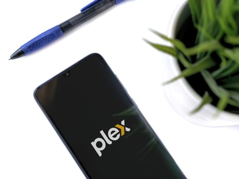 Lod, Israel - July 16,2023: Modern workspace with smartphone with Plex app launch screen on white background. Close up top view flat lay.