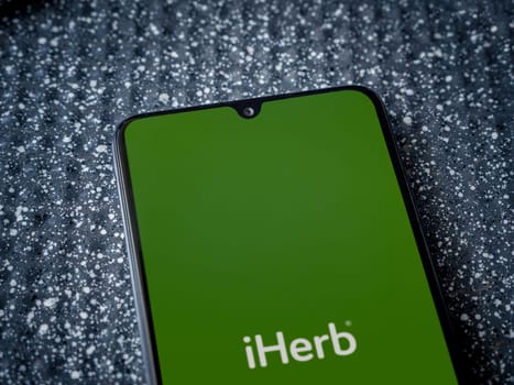 Lod, Israel - July 16,2023: iHerb app launch screen on the display of a black mobile smartphone on a metallic background. Close up top view flat lay.