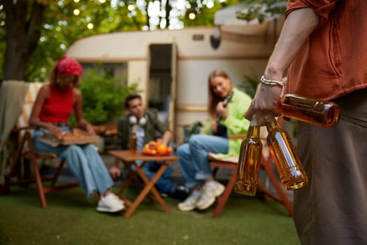 Group of happy friends resting in camping nearby their motorhome in forest. Focus on male hand holding several bottles of freshly cooled beer