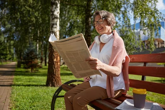 Happy smiling old woman sitting on wooden bench and reading newspaper relaxing outdoors in park