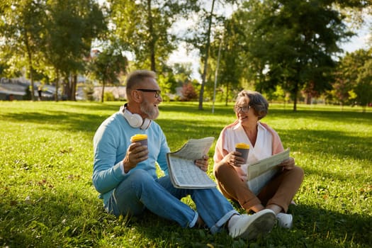 Senior loving couple drinking coffee and reading newspaper while relaxing on green lawn grass outdoors in park