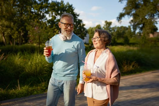 Positive elderly couple with refreshment cocktail walking outdoors over public green park background. Leisure activity on pension, carefree life of retirement